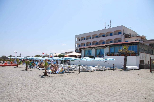 Facciata di un hotel fronte mare con ombrelloni e persone sulla spiaggia.