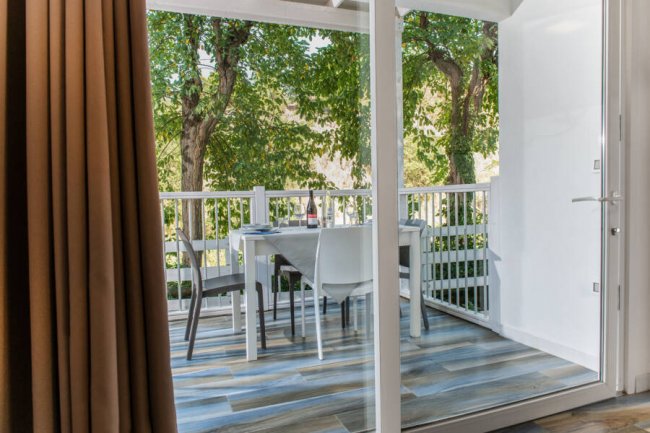 Balcone con tavolo apparecchiato, sedie bianche e vista sul giardino alberato.