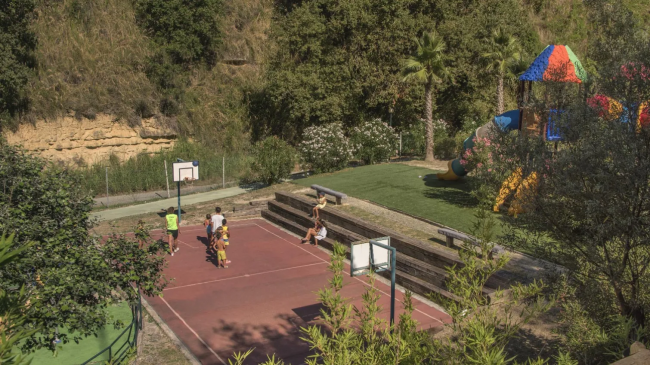 Campo da basket all'aperto accanto a un'area giochi per bambini, circondati da vegetazione.