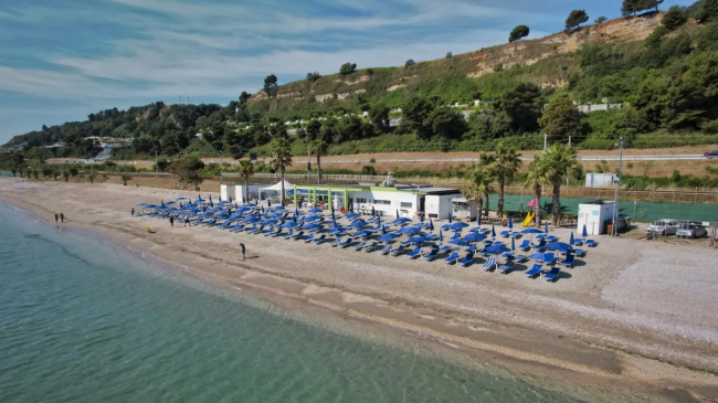 Spiaggia con file di ombrelloni blu, vicino a un tratto di strada e colline verdi.