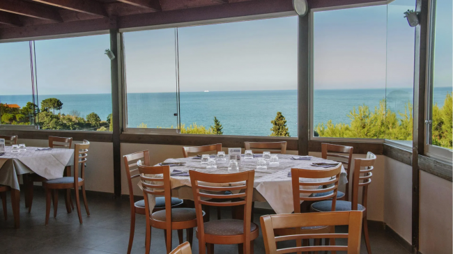 Ristorante con tavoli apparecchiati davanti a grandi finestre con vista sul mare e vegetazione.
