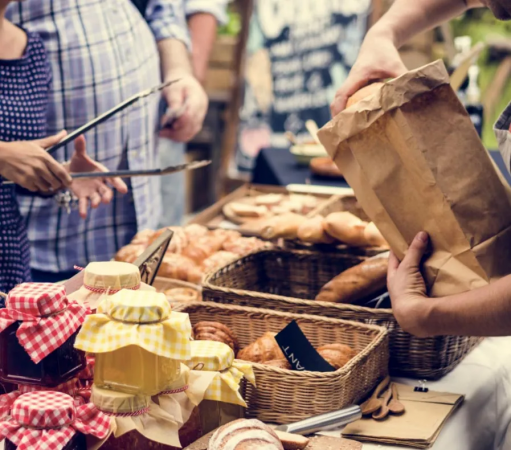 Persone scelgono prodotti da un tavolo con pane, dolci e marmellate in un mercato all'aperto.