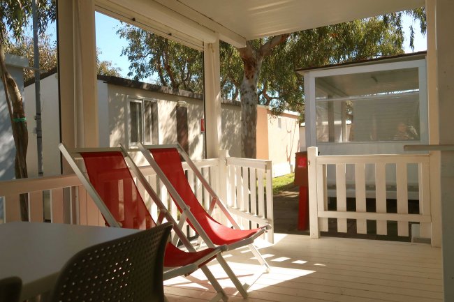 Veranda coperta con due sdraio rosse e tavolino, circondata da alberi e altre casette.