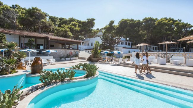 Piscina all'aperto con vegetazione, edifici bianchi e ombrelloni.