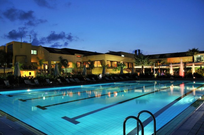 Piscina illuminata di sera con edificio sullo sfondo.