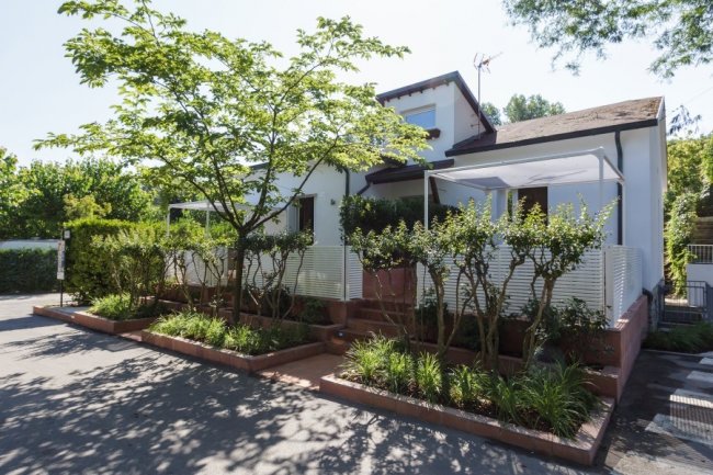 Casa bianca con terrazze, circondata da siepi e alberi.