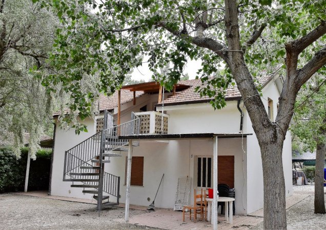 Casa bianca a due piani con scala esterna e tavolo sotto gli alberi.