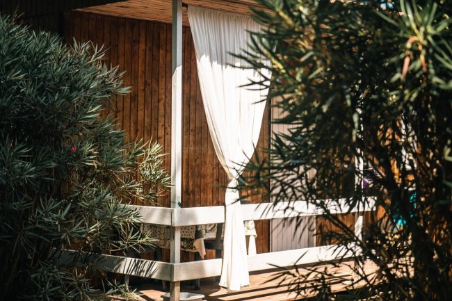Veranda in legno con tenda bianca tra piante verdi.