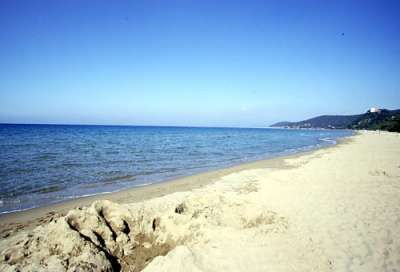 Spiaggia sabbiosa con mare calmo, cielo blu e colline in lontananza.