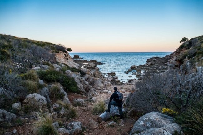 Uomo seduto su una roccia, guarda il mare tra scogli.