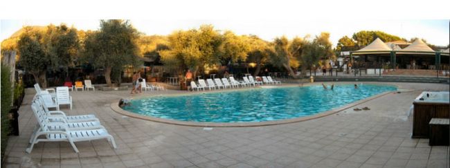 Una piscina all'aperto circondata da lettini, con alberi intorno e persone che nuotano.