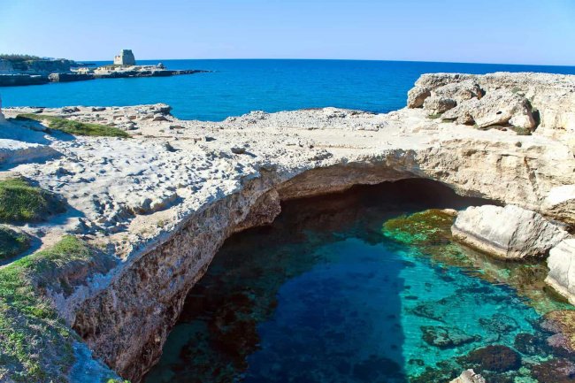 Acque limpide sotto una scogliera rocciosa, con torre lontana e mare blu.