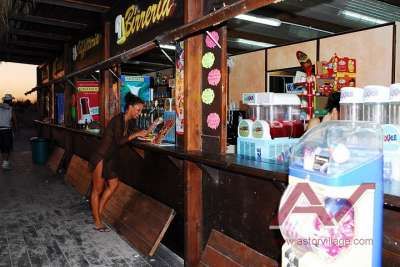 Bancone di uno stabilimento balneare, donna ordina una bevanda, luci calde e bevande esposte.