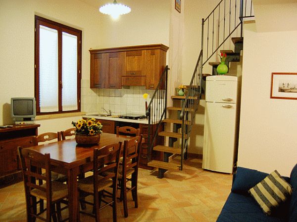 Interno di un appartamento con cucina, tavolo in legno, scale, divano e TV.