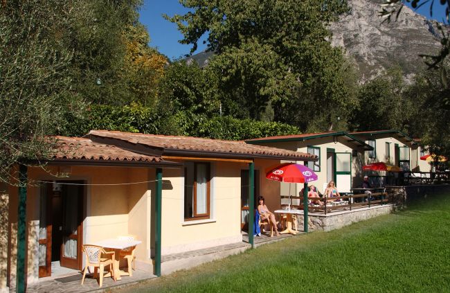 Bungalow in un campeggio, con tavolini all'aperto e ombrelloni colorati, circondati da alberi.