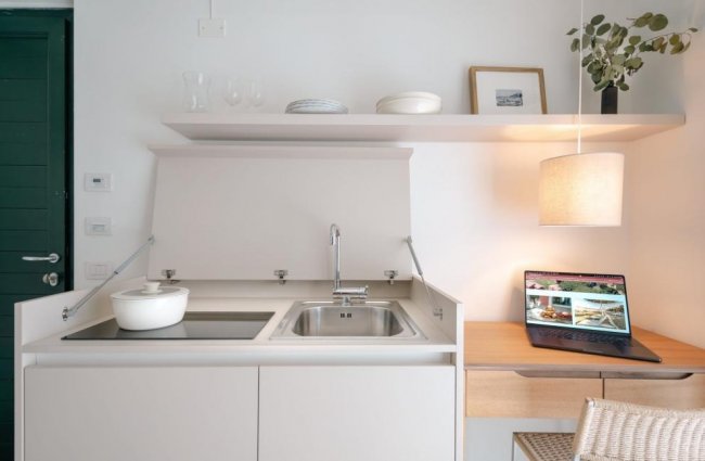 Cucina compatta con lavello, laptop e pentola bianca.