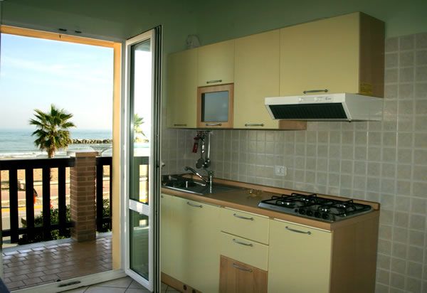 Cucina vista mare con mobili chiari, fornelli e cappa. Porta aperta verso balcone.