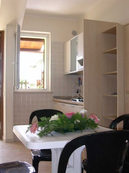 Cucina luminosa con tavolo bianco, sedie nere e fiori. Finestre aperte.