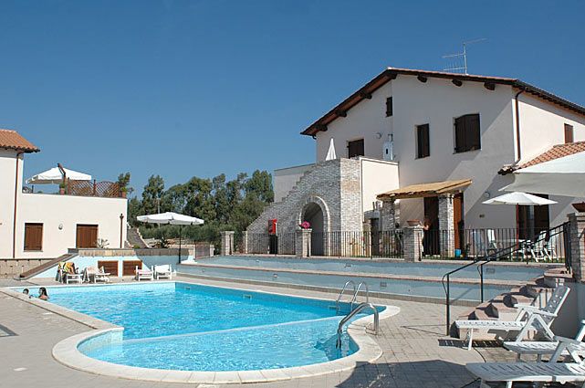 Piscina azzurra con sdraio vicino a edifici bianchi e tetti rossi sotto un cielo sereno.