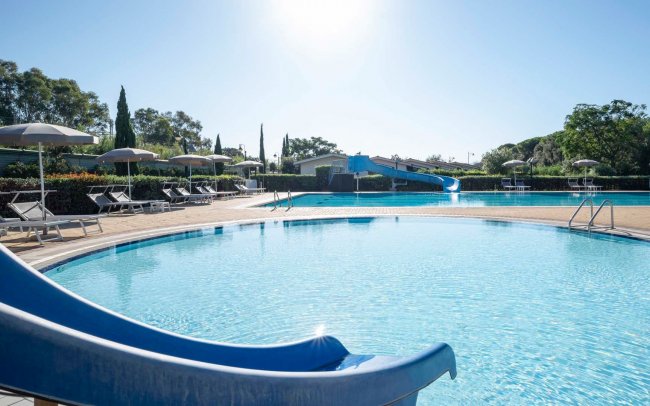 Piscina rotonda con scivolo blu e ombrelloni attorno, illuminata dal sole.