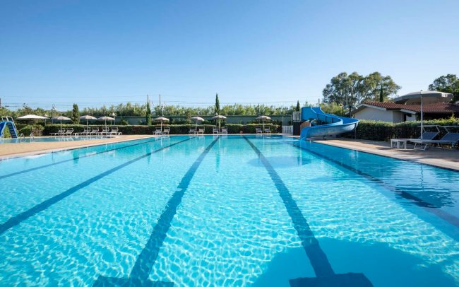 Piscina rettangolare con corsie, scivolo blu e lettini sotto ombrelloni.