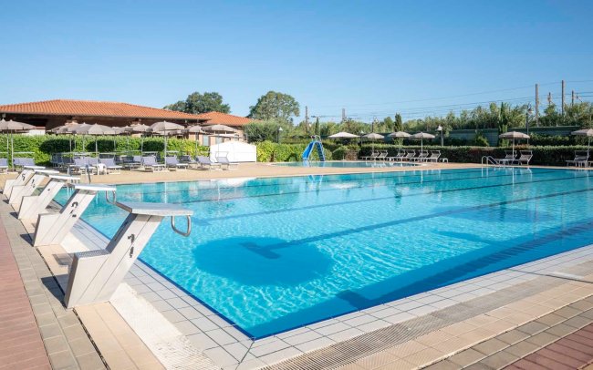 Piscina con trampolini, ombrelloni grigi e struttura con tetto rosso sullo sfondo.