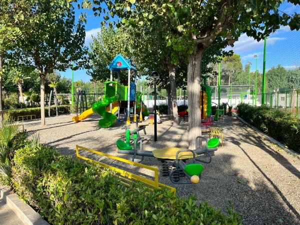 Un parco giochi con scivoli colorati e una giostrina, circondato da alberi verdi.
