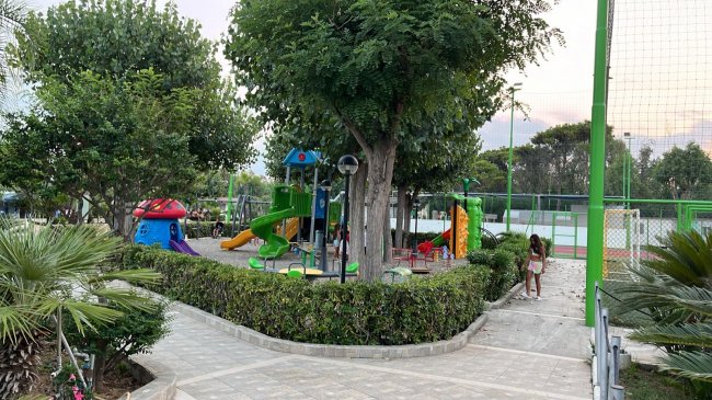 Un parco giochi con scivoli verdi e strutture colorate, circondato da alberi e un vialetto.