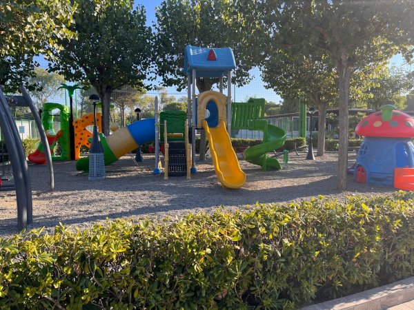 Un parco giochi con scivoli gialli e verdi, un'area in ghiaia e alberi intorno.
