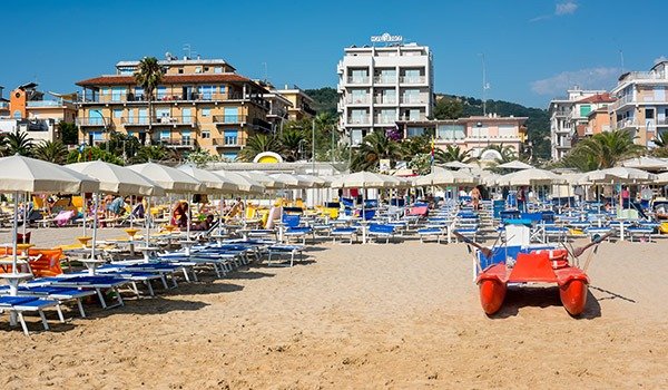 Spiaggia con ombrelloni e lettini, palazzi sullo sfondo.