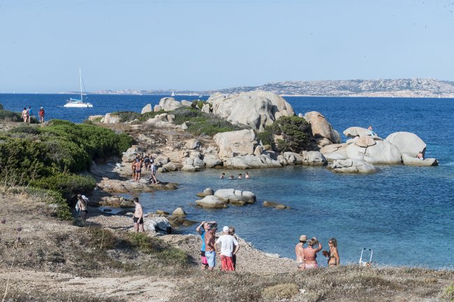 Persone su una spiaggia rocciosa con mare limpido e barca sullo sfondo.