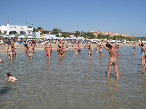 Persone che fanno esercizio in acqua poco profonda su una spiaggia affollata.
