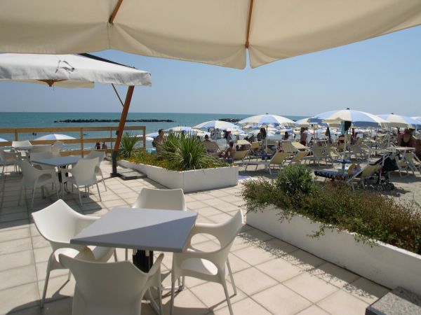 Terrazza esterna con tavoli sotto ombrelloni, vista mare e lettini sulla spiaggia.