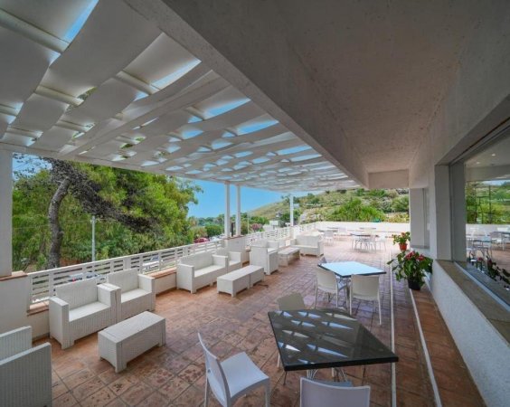Patio esterno con divani bianchi e tavoli, vista su alberi e colline.