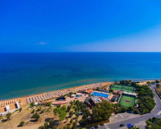 Vista aerea di un resort sulla spiaggia con ombrelloni, piscina e campi sportivi accanto al mare.