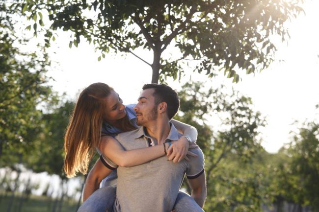 Coppia giovane, la donna sulla schiena dell'uomo, in un parco con alberi verdi.
