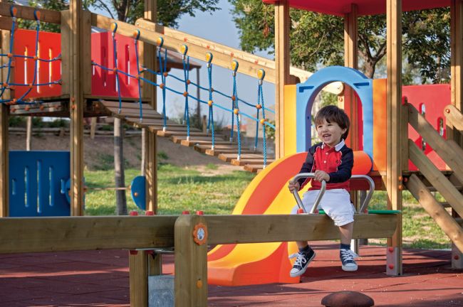 Bambino sorridente su un'altalena in legno, con scivolo arancione sullo sfondo.