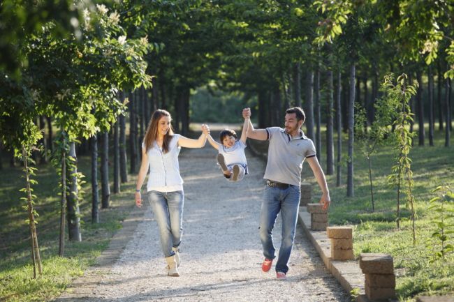 Tre persone camminano felici in un viale alberato, tenendo sollevato un bambino.