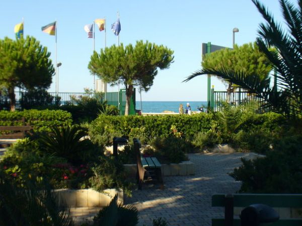 Giardino con panchine e palme, vista sul mare e bandiere sventolanti.