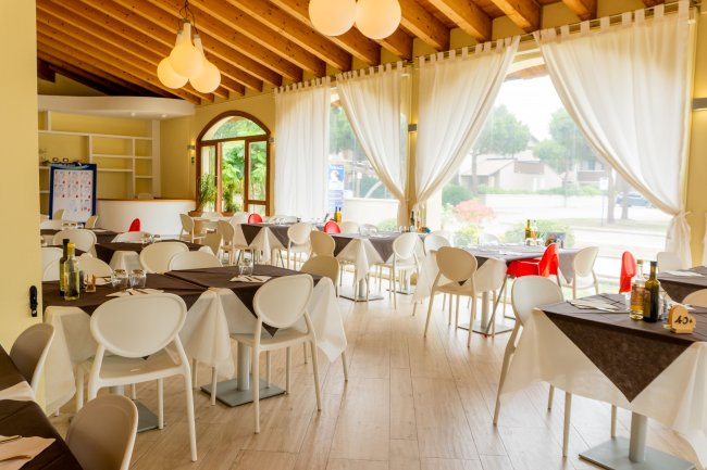 Sala ristorante luminosa con tavoli apparecchiati e tende bianche.