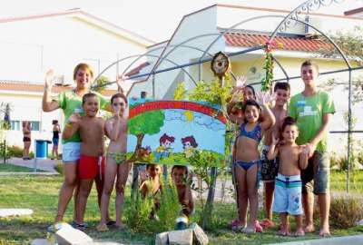 Un gruppo di bambini e adulti sorride davanti a un cartello colorato all'aperto.