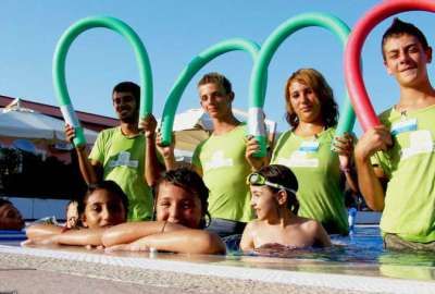 Bambini e adulti in piscina tengono cerchi colorati sopra la testa e sorridono.