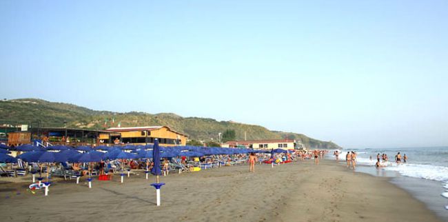 Spiaggia con ombrelloni blu, bagnanti sulla sabbia e mare.
