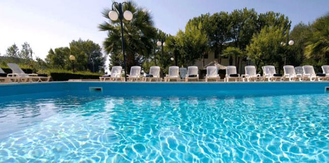 Piscina azzurra circondata da lettini bianchi e alberi.