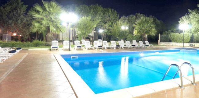 Piscina illuminata di notte con lettini e alberi.
