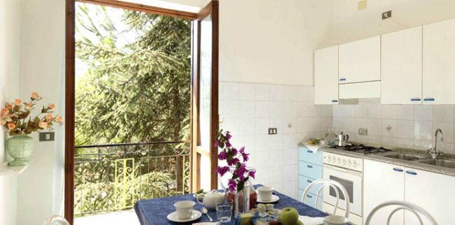 Cucina luminosa con tavolo apparecchiato, finestre aperte su alberi esterni.