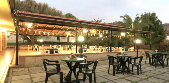 Terrazza coperta con tavoli e sedie in plastica, decorata con festoni, immersa nel verde.