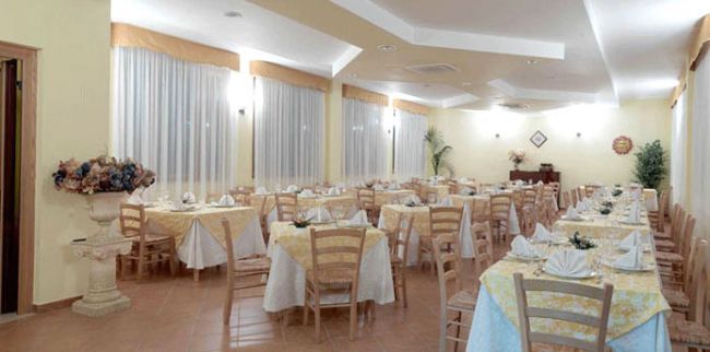 Sala da pranzo luminosa con tavoli apparecchiati, sedie di legno e tende bianche alle finestre.