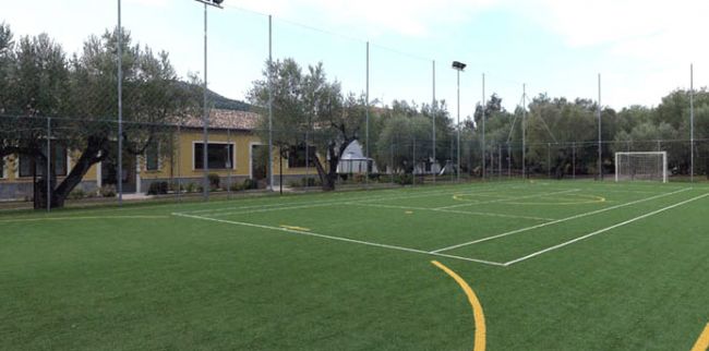 Un campo da calcio con erba sintetica circondato da alberi e un edificio giallo.