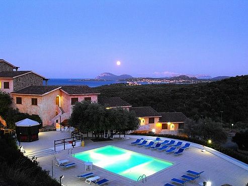 Piscina illuminata al tramonto con vista sul mare e colline in lontananza.
